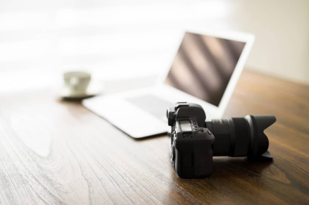 Professional DSLR camera on wooden table with open laptop and cup of coffee in background