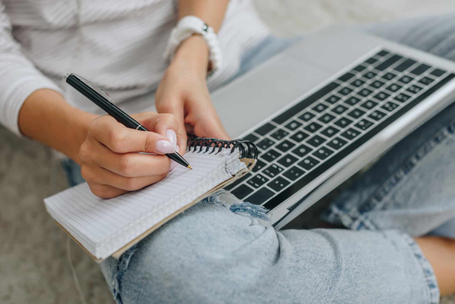 Woman writing in notebook with laptop on lap
